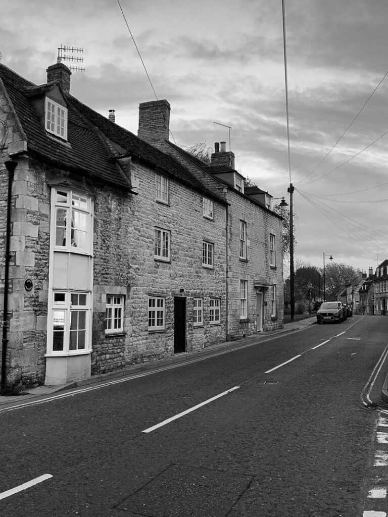 A black and white photo of a quiet street scene featuring stone buildings on either side, with a visible road and overcast sky.